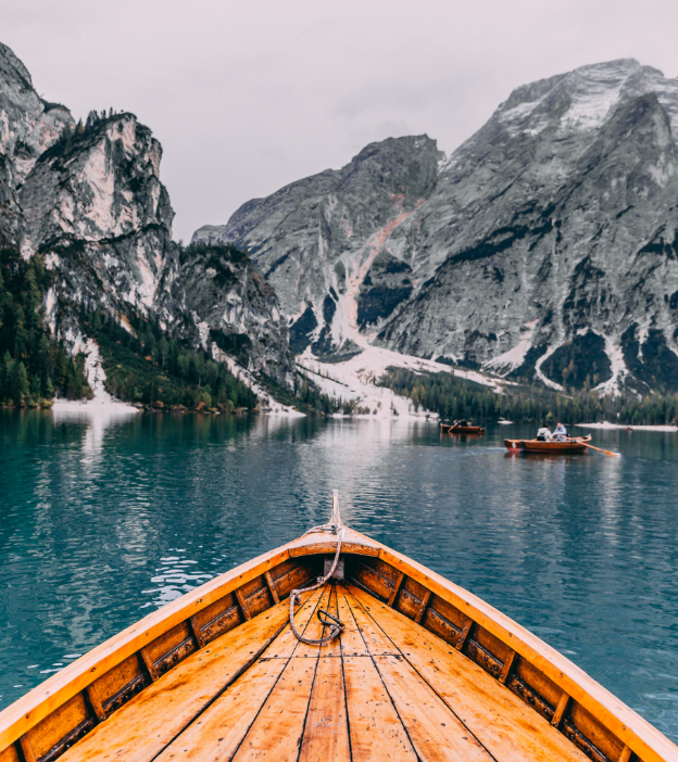 The bow of a wood panelled boat heads into a bay of water, surrounded by rocky mountains