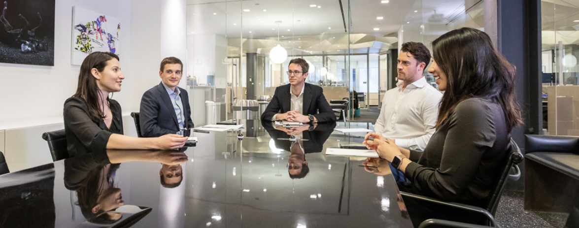 Five colleagues sit around a long table in a conference room with glass walls.